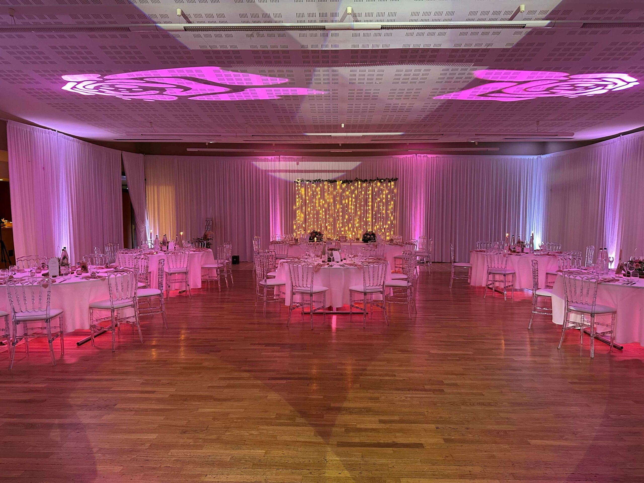 Aménagement de salle de mariage, rideau au mur, table ronde lumineuse, chaise napoléon transparente et éclairage d'ambiance. Location Jani Event.